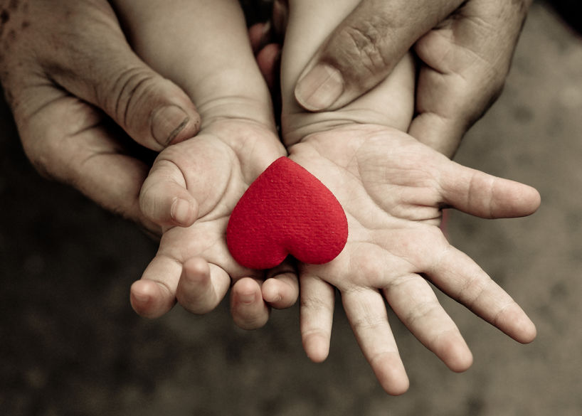 A young child's hands holding a red heart, supported by an older person's hands.